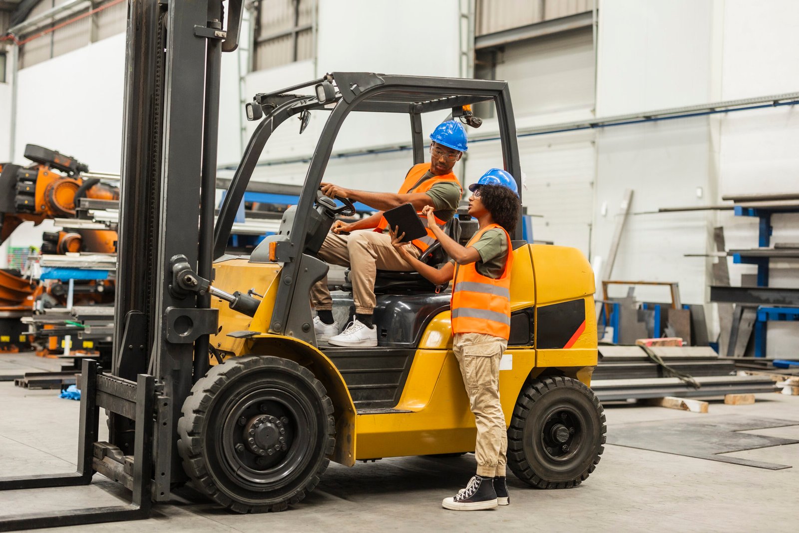 Engineers using digital tablet and driving forklift in factory Two multiracial industrial engineers wearing safety vests and helmets are using a digital tablet and driving a forklift truck inside a large factory